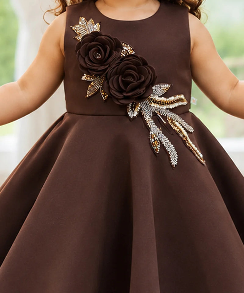 A sleeveless brown dress with a large dark brown fabric flower and gold and silver embellished leaves on the bodice, worn by a child.