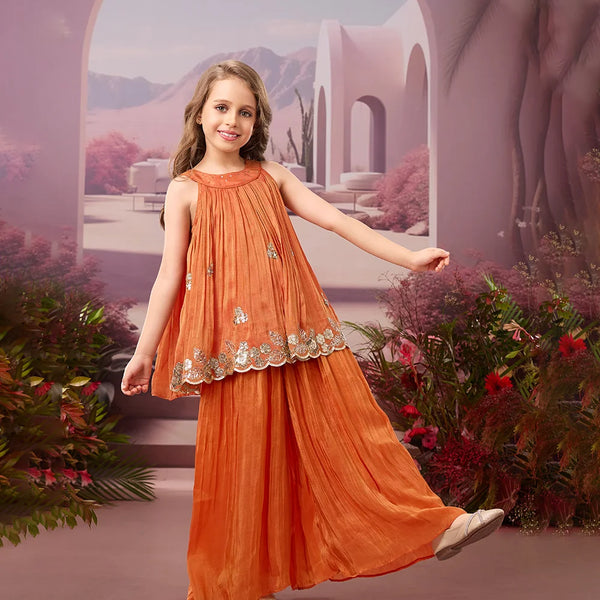 Young girl in an orange traditional outfit standing in a decorative setting with arches and plants.