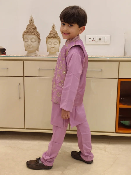 It is a side profile look of the boy wearing a lavender color kurta pajama with a gold thread embroidery.
