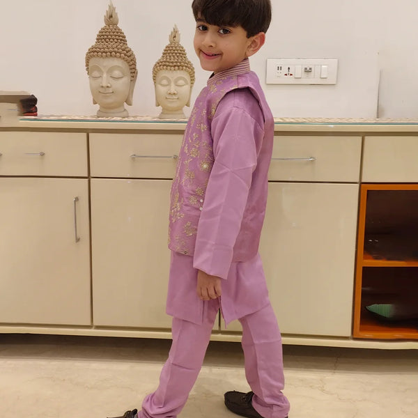 It is a side profile look of the boy wearing a lavender color kurta pajama with a gold thread embroidery.