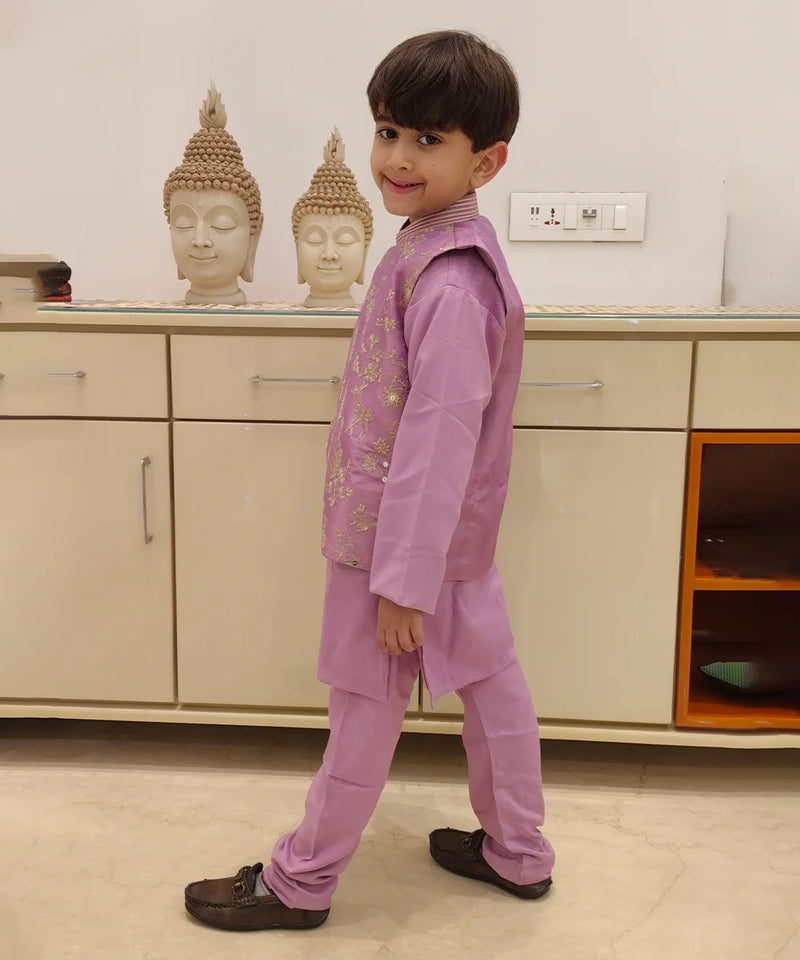 It is a side profile look of the boy wearing a lavender color kurta pajama with a gold thread embroidery.