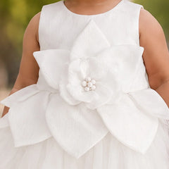 A close-up of a child wearing a sleeveless white dress with a large layered flower applique at the waist and a tulle skirt, photographed outdoors against a blurred green background.
