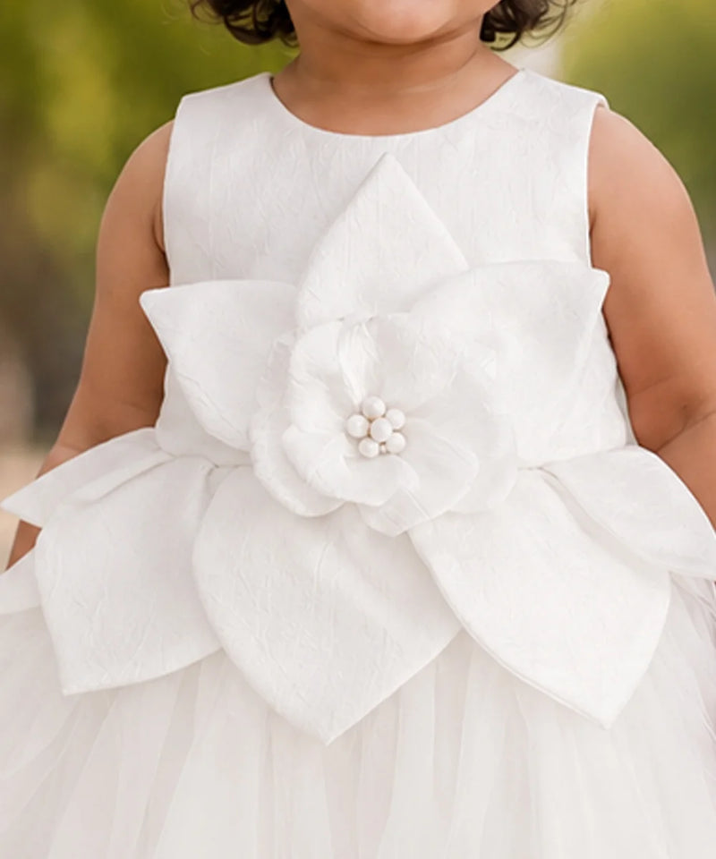 A close-up of a child wearing a sleeveless white dress with a large layered flower applique at the waist and a tulle skirt, photographed outdoors against a blurred green background.