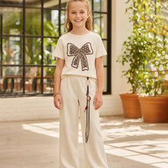 A young girl stands in a bright indoor-outdoor space wearing a cream short-sleeve t-shirt with a large black sequined bow graphic on the front, paired with matching cream wide-leg pants and white shoes.