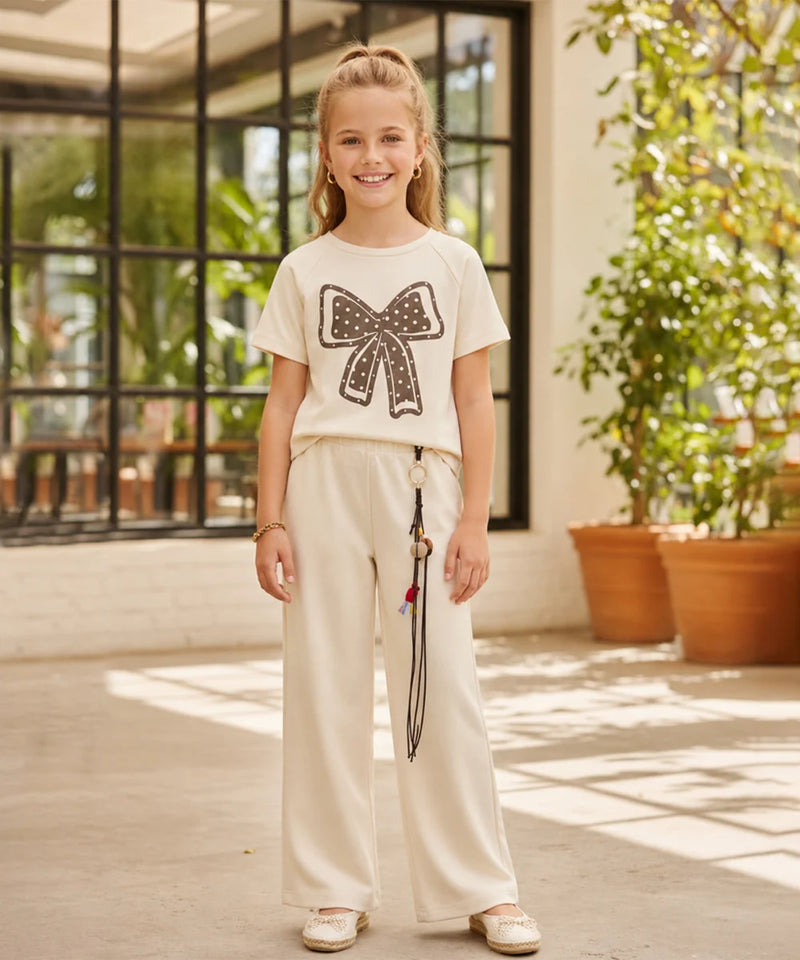 A young girl stands in a bright indoor-outdoor space wearing a cream short-sleeve t-shirt with a large black sequined bow graphic on the front, paired with matching cream wide-leg pants and white shoes.