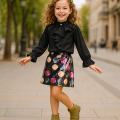 A young girl with curly hair stands on a city sidewalk wearing a black ruffled long-sleeve blouse and a black sequined skirt with colorful floral patterns, paired with olive green ankle boots.