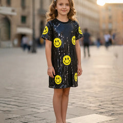 A young girl stands in a cobblestone street wearing a short-sleeve black sequined dress decorated with yellow smiley face prints, paired with white shoes.