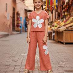 A young girl with braided hair stands on a cobblestone street wearing a terracotta-colored two-piece set with large white flower appliqués, paired with tan sandals. The background shows a blurred outdoor market scene with buildings and colorful flags.