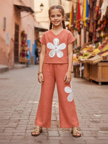 A young girl with braided hair stands on a cobblestone street wearing a terracotta-colored two-piece set with large white flower appliqués, paired with tan sandals. The background shows a blurred outdoor market scene with buildings and colorful flags.