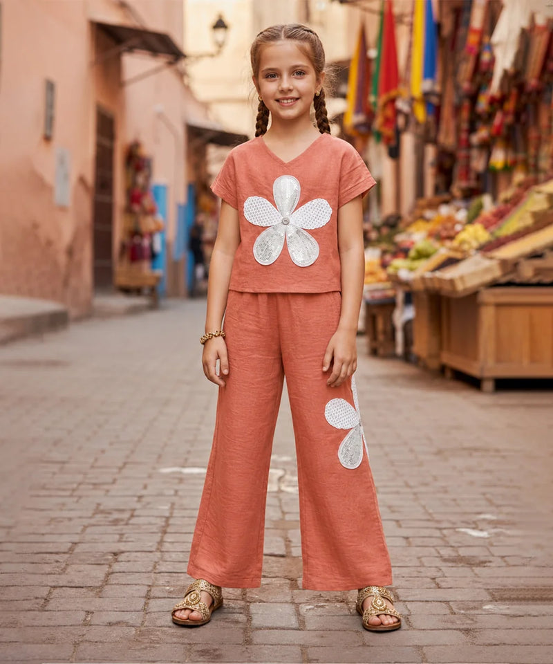 A young girl with braided hair stands on a cobblestone street wearing a terracotta-colored two-piece set with large white flower appliqués, paired with tan sandals. The background shows a blurred outdoor market scene with buildings and colorful flags.