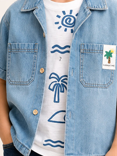 A close-up of a person wearing a light blue denim short-sleeve button-up shirt over a white graphic T-shirt with blue sun, wave, palm tree, and beach hat illustrations. A small white chest patch on the denim reads 'SUMMER TIME' with a palm tree.