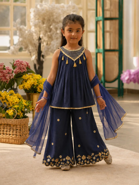Young girl in a traditional navy blue outfit with gold details standing in a decorated room.