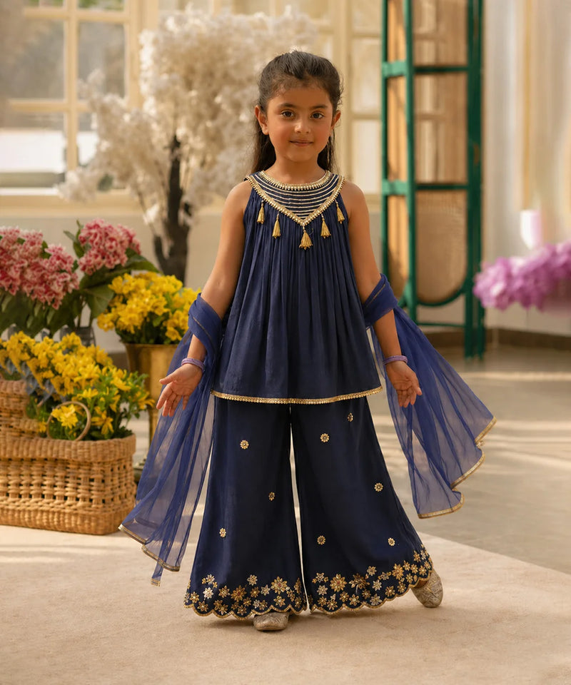 Young girl in a traditional navy blue outfit with gold details standing in a decorated room.