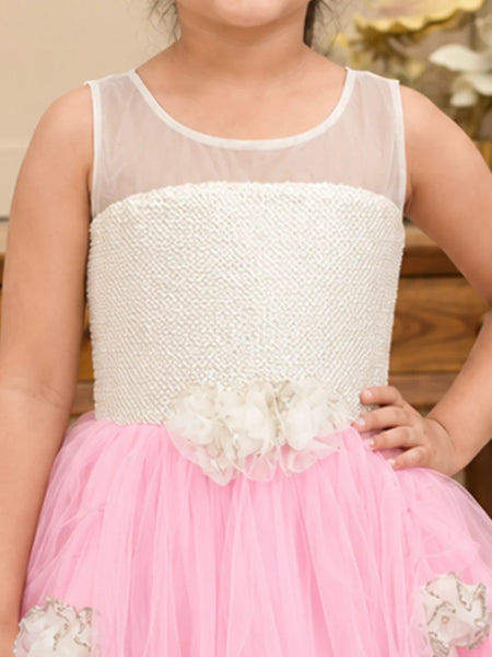 A young girl wearing a sleeveless white and pink flower girl dress with a beaded bodice, sheer neckline, and a pink tulle skirt with white fabric flowers.