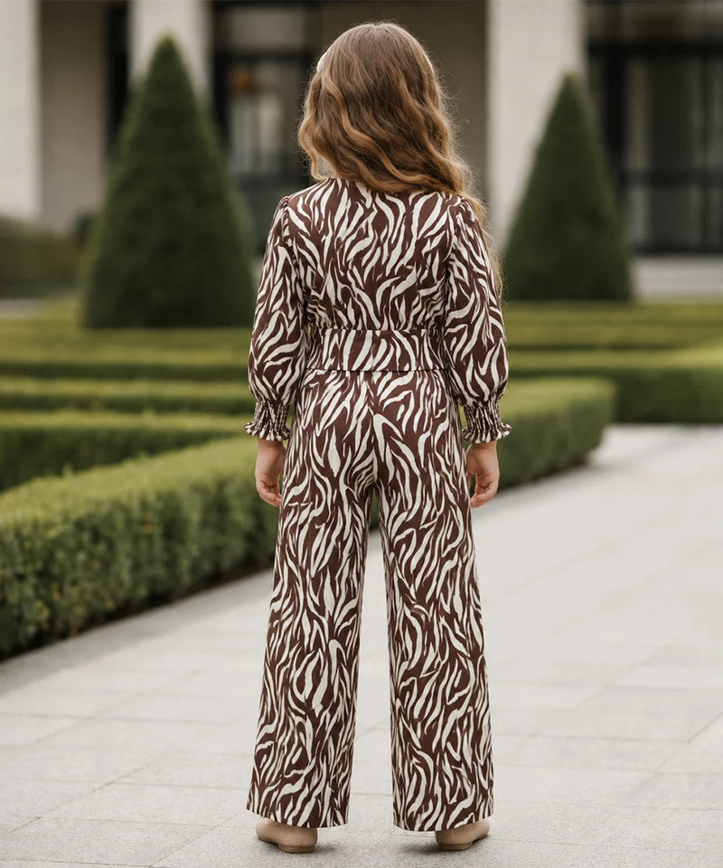A woman viewed from behind wearing a brown and white zebra-print jumpsuit, standing on a paved walkway in front of manicured hedges and trees.