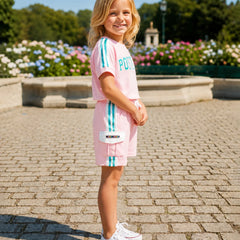 A young girl stands outdoors on a cobblestone path wearing a light pink two-piece tracksuit with turquoise stripes and white sneakers. The background shows a park with flowers and trees.