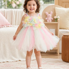 A young girl with curly brown hair stands in a cozy, sunlit room wearing a pastel rainbow flower and tulle tutu dress. The dress features a floral bodice with pastel yellow, green, blue, and pink flowers, supported by thin pastel rainbow straps, and a layered tulle skirt in pink, white, and light blue.