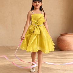 A young girl with long dark hair wearing a bright yellow sleeveless party dress with a large bow at the waist, white shoes, and a small purple bow in her hair, holding a pink and blue ribbon. She is standing in front of a beige wall with a terracotta pot in the background.