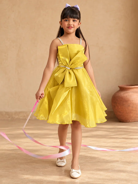 A young girl with long dark hair wearing a bright yellow sleeveless party dress with a large bow at the waist, white shoes, and a small purple bow in her hair, holding a pink and blue ribbon. She is standing in front of a beige wall with a terracotta pot in the background.