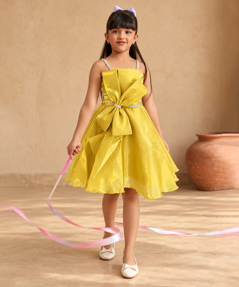 A young girl with long dark hair wearing a bright yellow sleeveless party dress with a large bow at the waist, white shoes, and a small purple bow in her hair, holding a pink and blue ribbon. She is standing in front of a beige wall with a terracotta pot in the background.