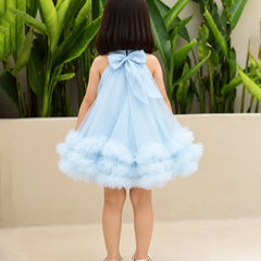 A young girl viewed from behind wearing a light blue halter dress with a large bow at the back and a fluffy tulle skirt, standing outdoors in front of green plants and a beige wall.