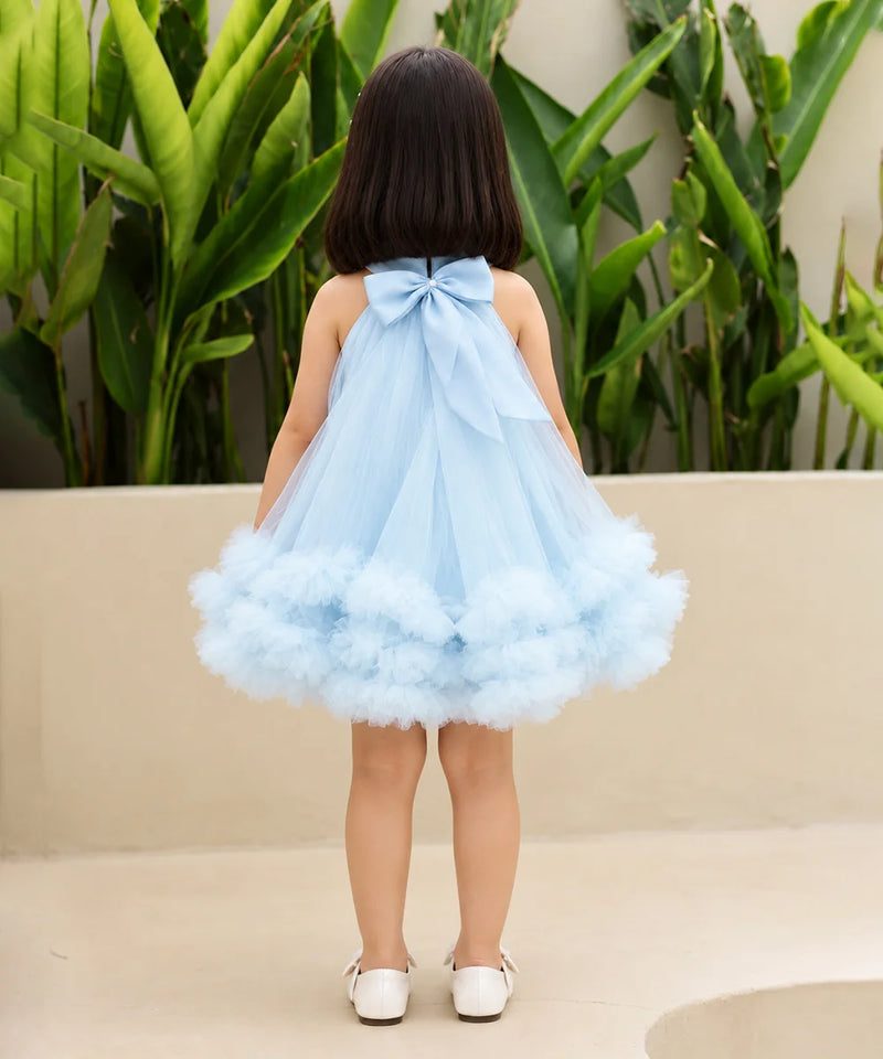 A young girl viewed from behind wearing a light blue halter dress with a large bow at the back and a fluffy tulle skirt, standing outdoors in front of green plants and a beige wall.