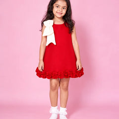 A young girl with long dark hair stands against a solid pink background wearing a red sleeveless dress with a large cream bow on the shoulder and a ruffled hem, paired with white ruffled socks.
