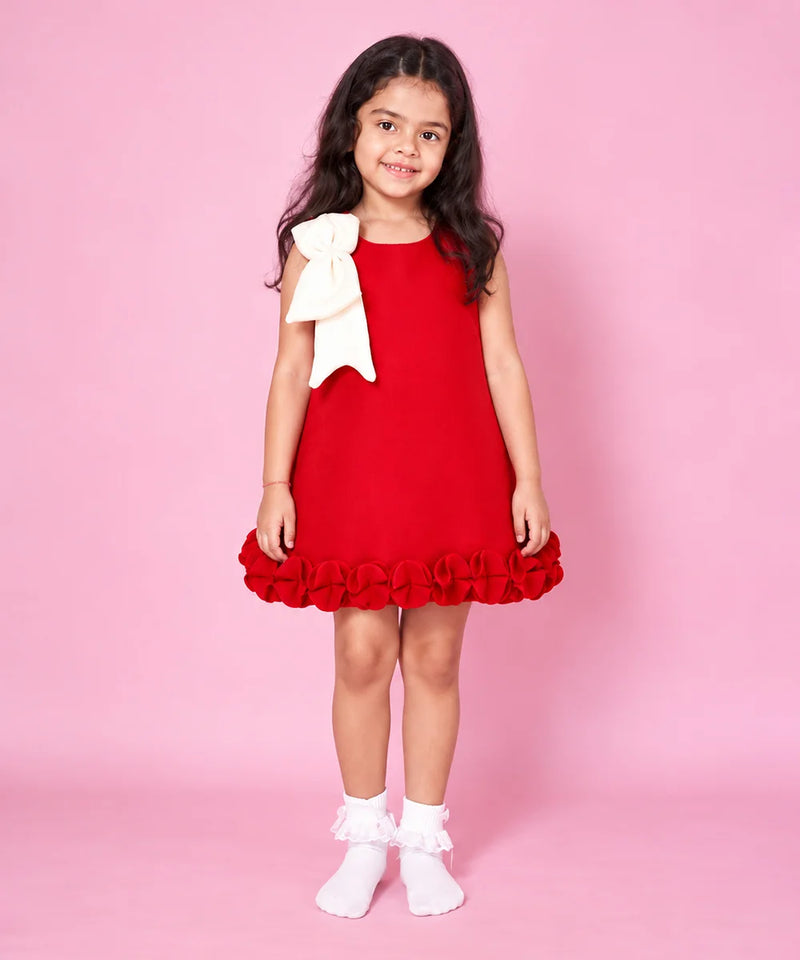 A young girl with long dark hair stands against a solid pink background wearing a red sleeveless dress with a large cream bow on the shoulder and a ruffled hem, paired with white ruffled socks.