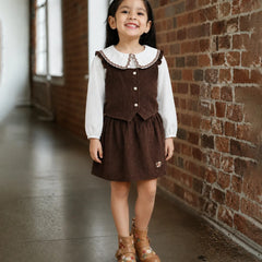 A young girl stands in a brick hallway wearing a brown corduroy pinafore-style dress over a white long-sleeve shirt, paired with tan lace-up ankle boots.