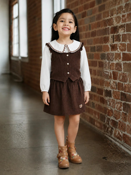 A young girl stands in a brick hallway wearing a brown corduroy pinafore-style dress over a white long-sleeve shirt, paired with tan lace-up ankle boots.