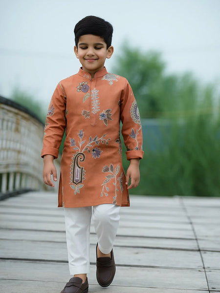 Child wearing an embroidered orange kurta with white pants on a wooden deck.