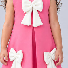 A sleeveless pink dress with large white bow details on the front and lower sides, shown on a child against a light gray background.