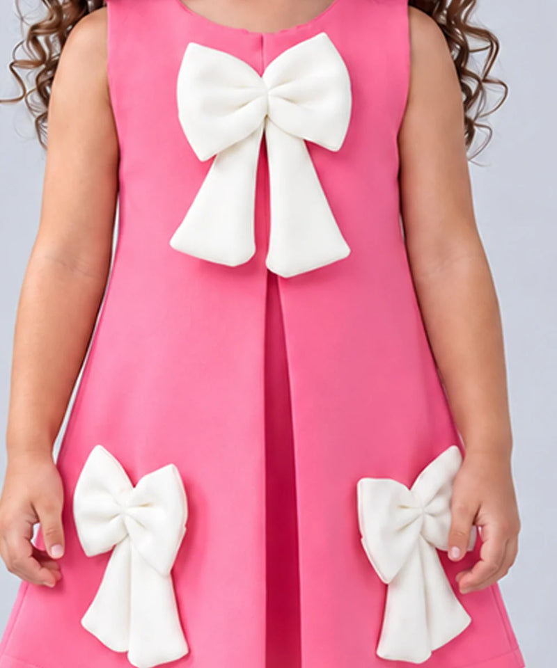 A sleeveless pink dress with large white bow details on the front and lower sides, shown on a child against a light gray background.