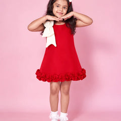 A young girl modeling a red sleeveless dress with a large white bow on the shoulder and a ruffled hem, standing against a solid pink background.