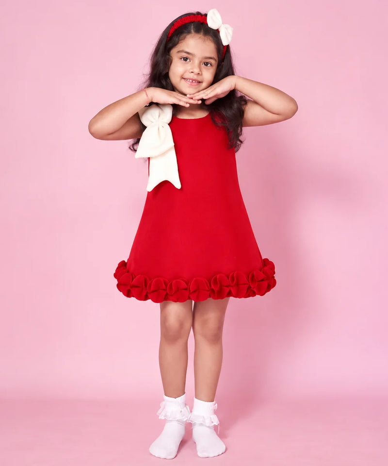 A young girl modeling a red sleeveless dress with a large white bow on the shoulder and a ruffled hem, standing against a solid pink background.