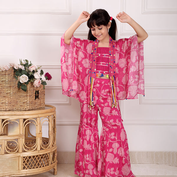 Child wearing a pink floral outfit with arms raised in a room with a wicker table and flowers.