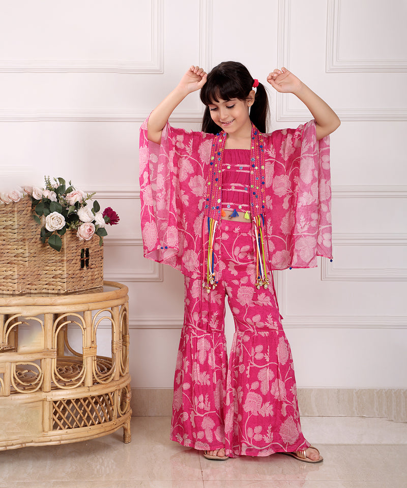 Child wearing a pink floral outfit with arms raised in a room with a wicker table and flowers.