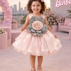 A young girl stands outdoors wearing a shiny light pink party dress with a large gray and pink flower applique at the chest. The background shows a pink-themed patio with balloons, flowers, a sofa, and a neon sign.