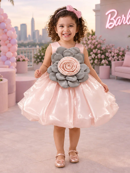 A young girl stands outdoors wearing a shiny light pink party dress with a large gray and pink flower applique at the chest. The background shows a pink-themed patio with balloons, flowers, a sofa, and a neon sign.