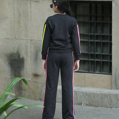 A person viewed from behind wearing a black tracksuit with bright pink and neon yellow side stripes, standing outdoors against a stone wall.