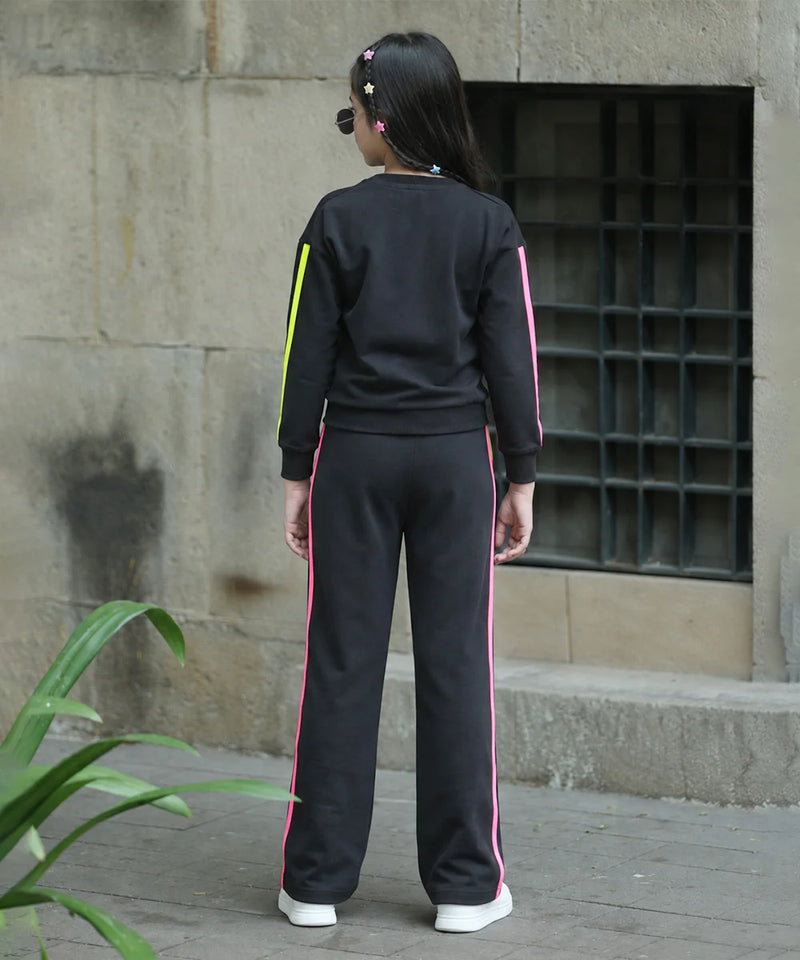 A person viewed from behind wearing a black tracksuit with bright pink and neon yellow side stripes, standing outdoors against a stone wall.