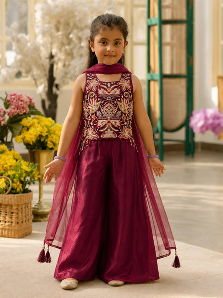 Young girl in a maroon traditional outfit with floral patterns standing in a decorated room.