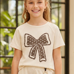 A young girl smiling while wearing a cream-colored short-sleeve t-shirt with a brown bow graphic on the front, paired with cream pants.