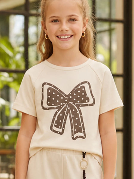 A young girl smiling while wearing a cream-colored short-sleeve t-shirt with a brown bow graphic on the front, paired with cream pants.