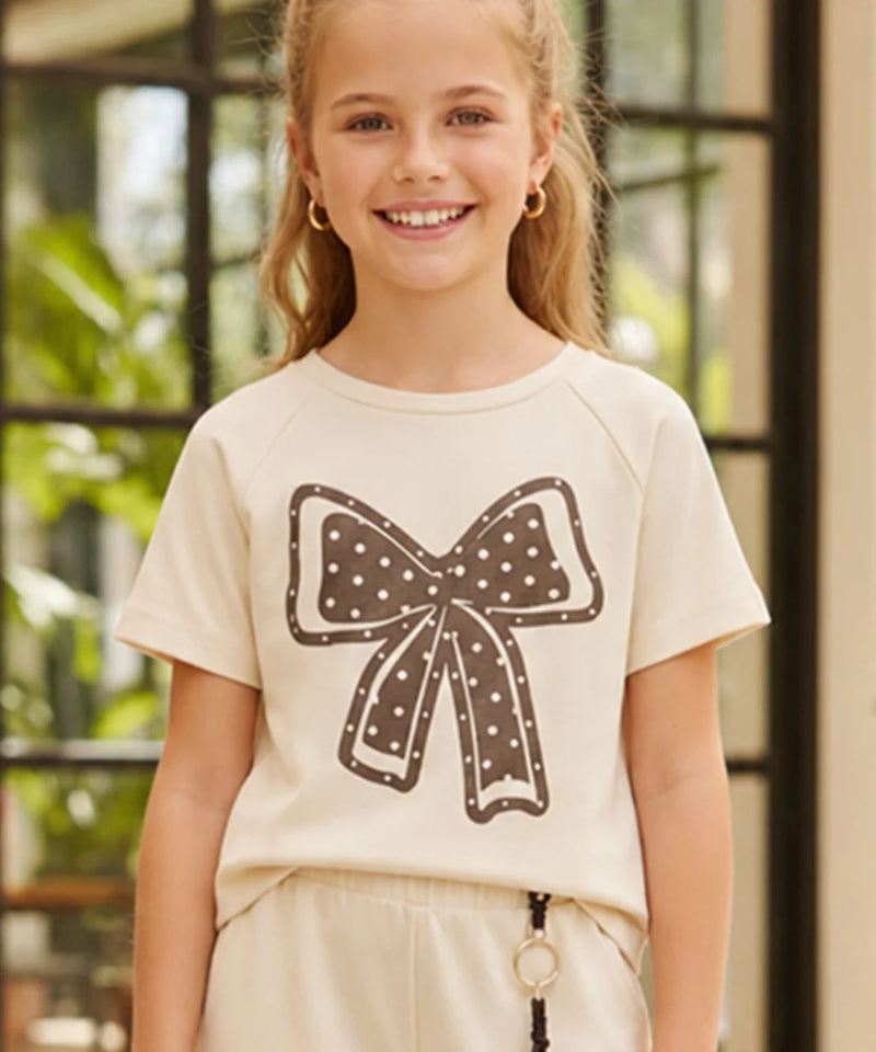 A young girl smiling while wearing a cream-colored short-sleeve t-shirt with a brown bow graphic on the front, paired with cream pants.