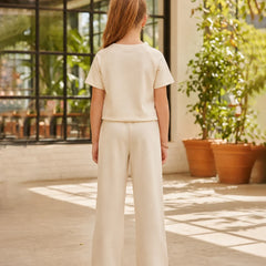 A child standing with their back to the camera, wearing a matching cream-colored short-sleeve top and wide-leg trousers set, in an outdoor patio setting with potted plants.