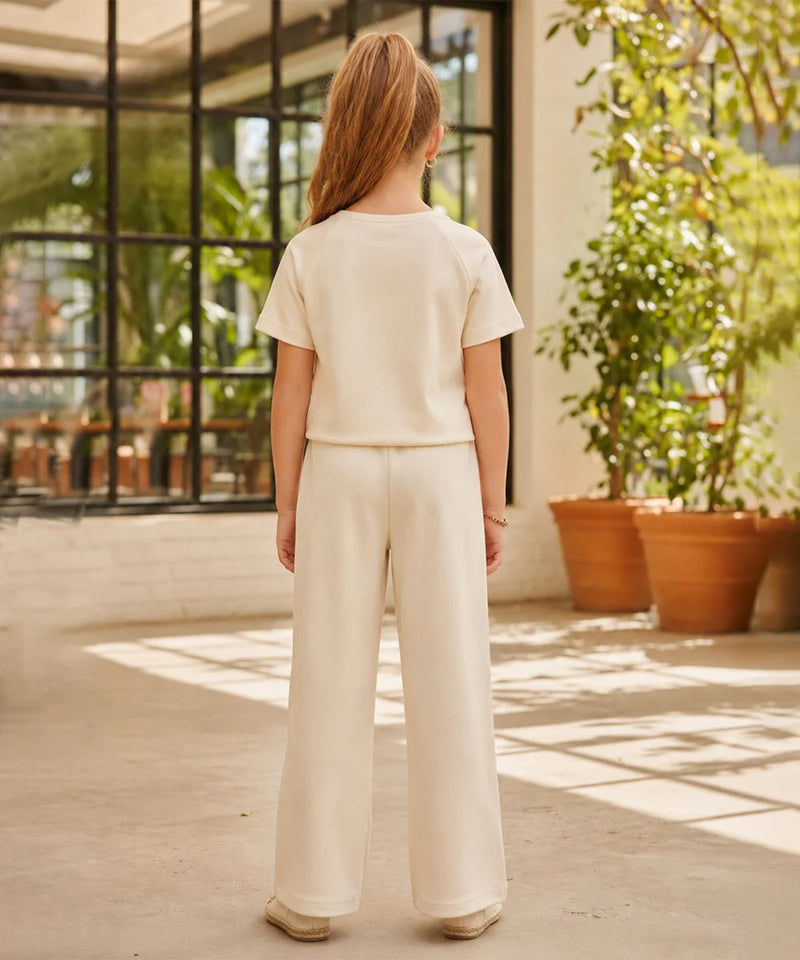 A child standing with their back to the camera, wearing a matching cream-colored short-sleeve top and wide-leg trousers set, in an outdoor patio setting with potted plants.