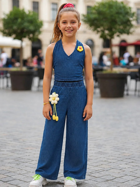 A young girl stands outdoors in a paved plaza wearing a sleeveless blue denim-like jumpsuit with wide legs, white sneakers, and yellow flower accessories.