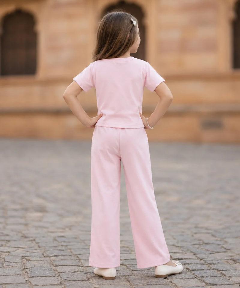 A young girl seen from behind wearing a light pink short-sleeve t-shirt and matching wide-leg trousers, standing on a cobblestone surface with a blurred stone building in the background.