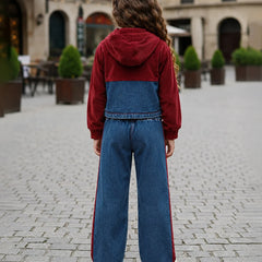 A child standing outdoors on a cobblestone street, viewed from behind, wearing a color-block hoodie with a burgundy top and hood, a blue denim middle panel, and matching wide-leg blue denim pants with red side stripes.